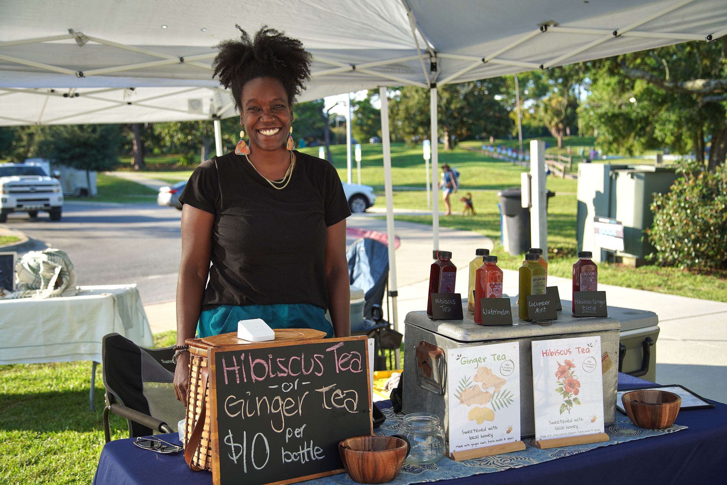 Woman smiling at outdoor market booth selling Hibiscus Tea and Ginger Tea for $10 per bottle.