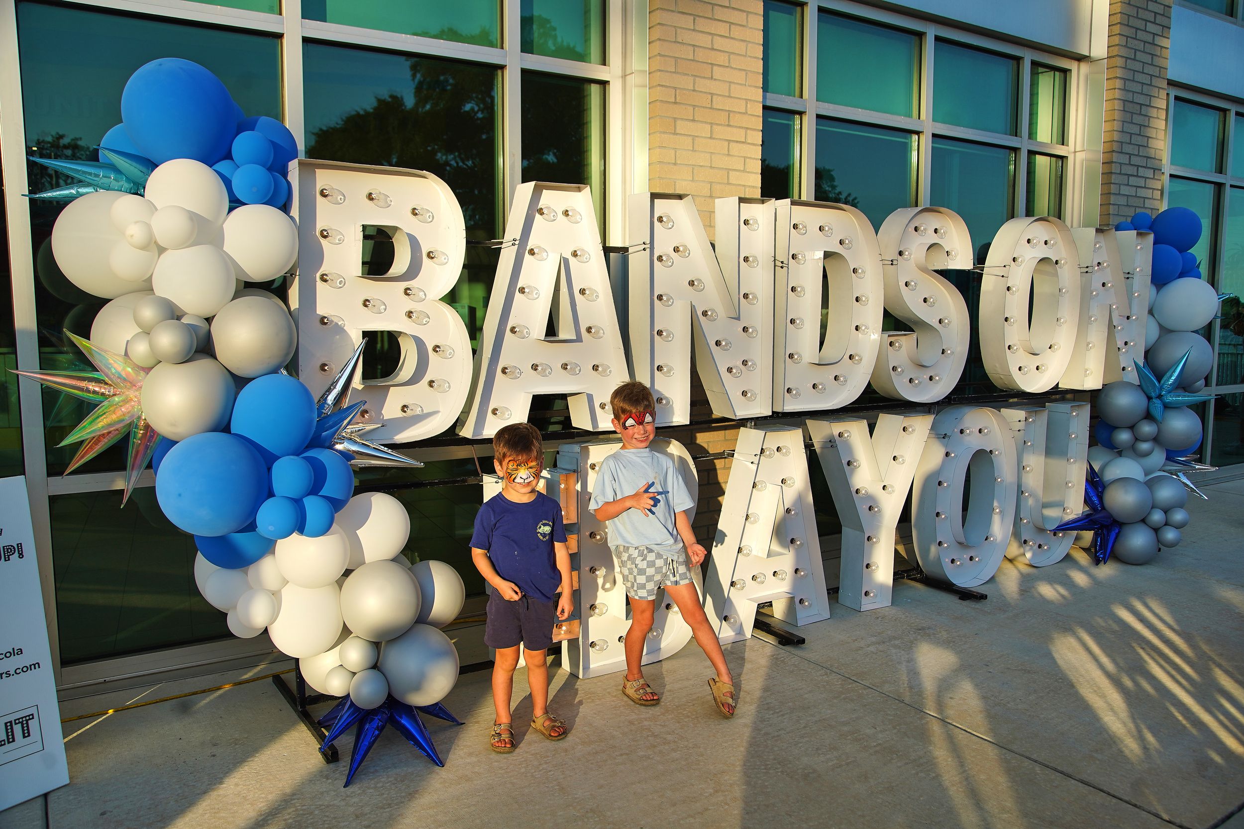 Two people pose in front of large marquee Bands on Bayou sign with blue and white balloon decorations outdoors.