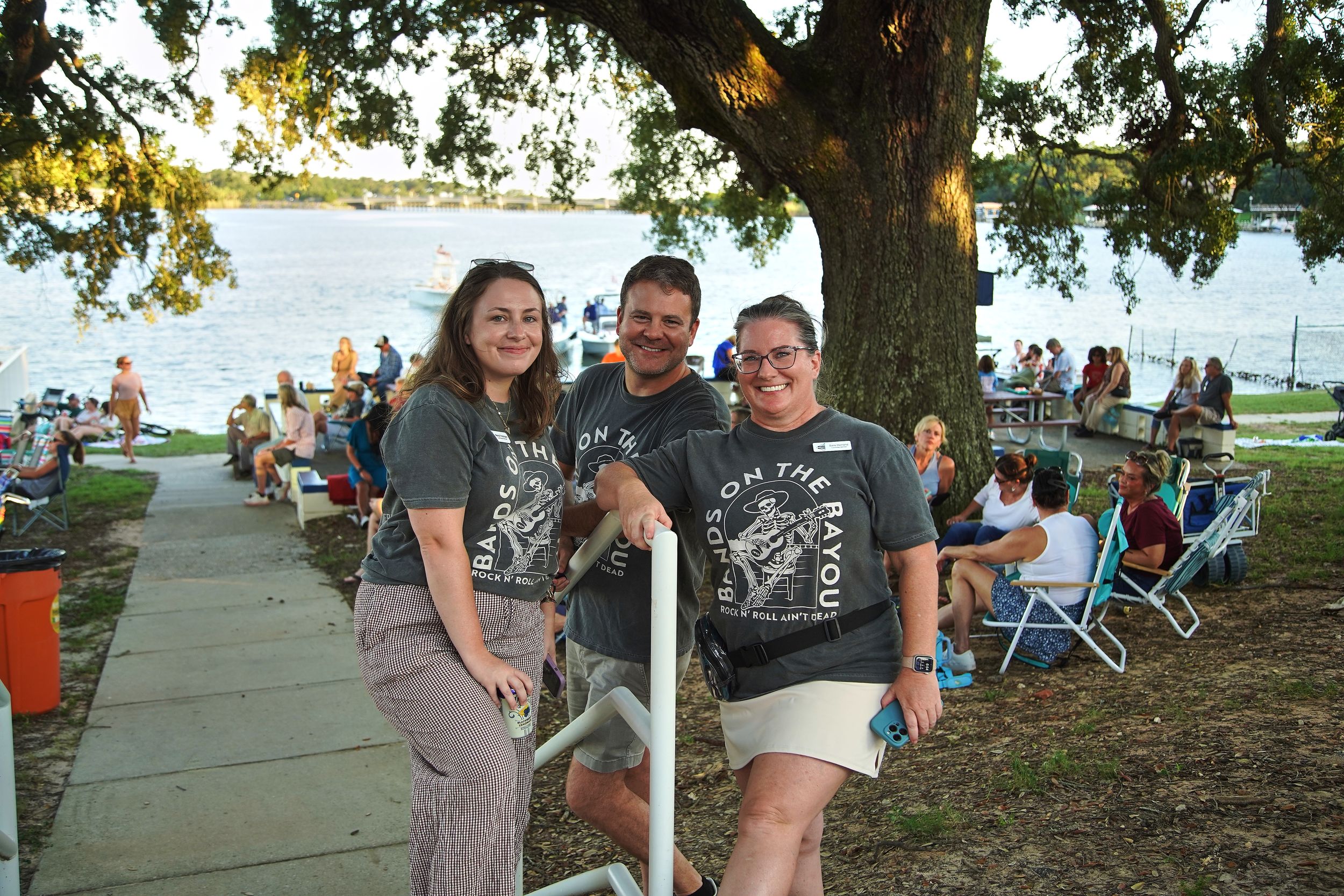 Three friends pose smiling near a lakeside park during an outdoor evening event with crowd in background.