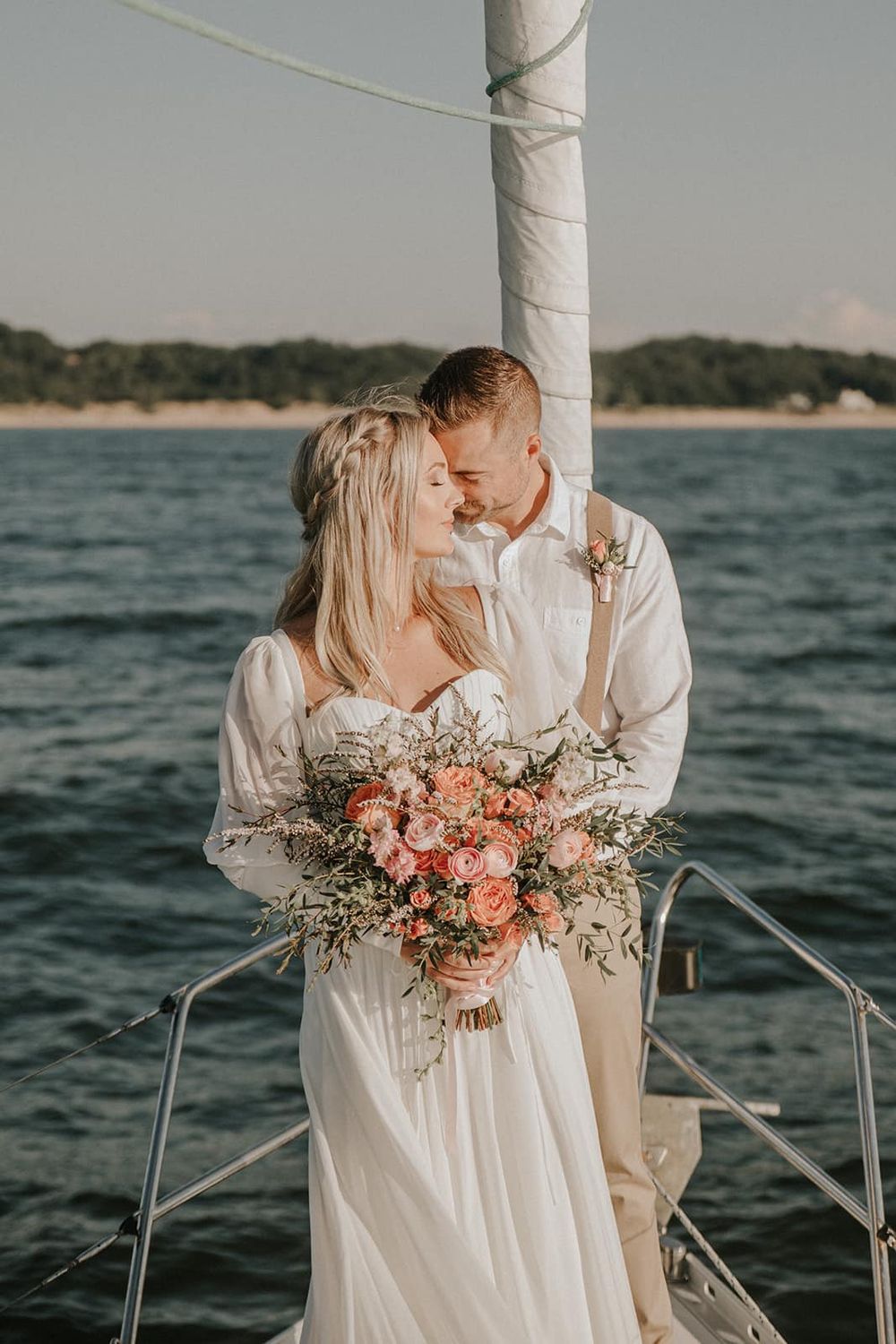 Bride and groom share a romantic moment on a sailboat, bride holding a pink bouquet with ocean backdrop.