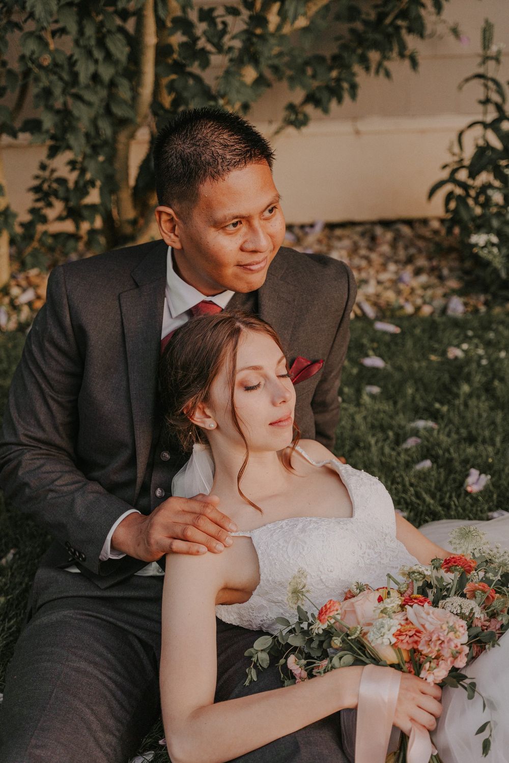 A bride and groom sit together outdoors, she rests on him holding a floral bouquet, both in elegant wedding attire.