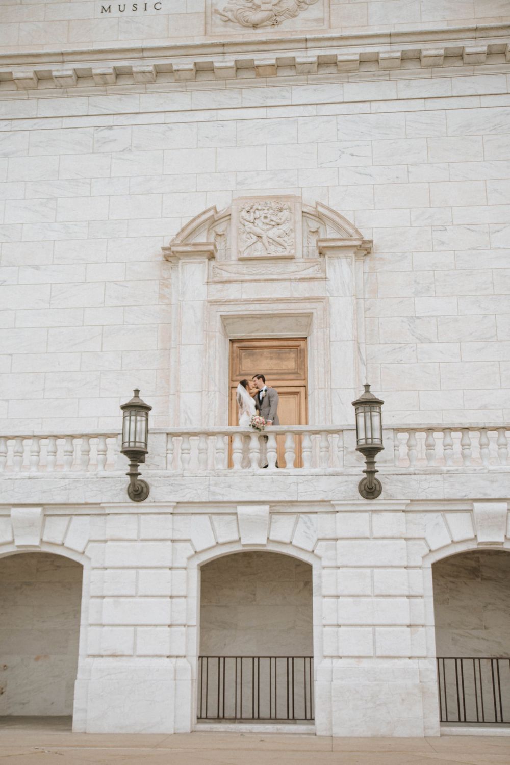 Couple sharing a romantic kiss on a grand white marble balcony of a classical music hall building.