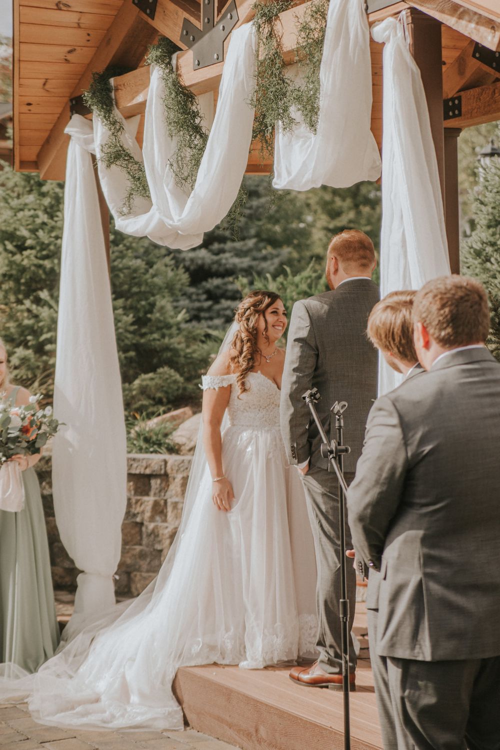 Bride in white off-shoulder gown stands at outdoor wedding altar adorned with white draping and greenery.