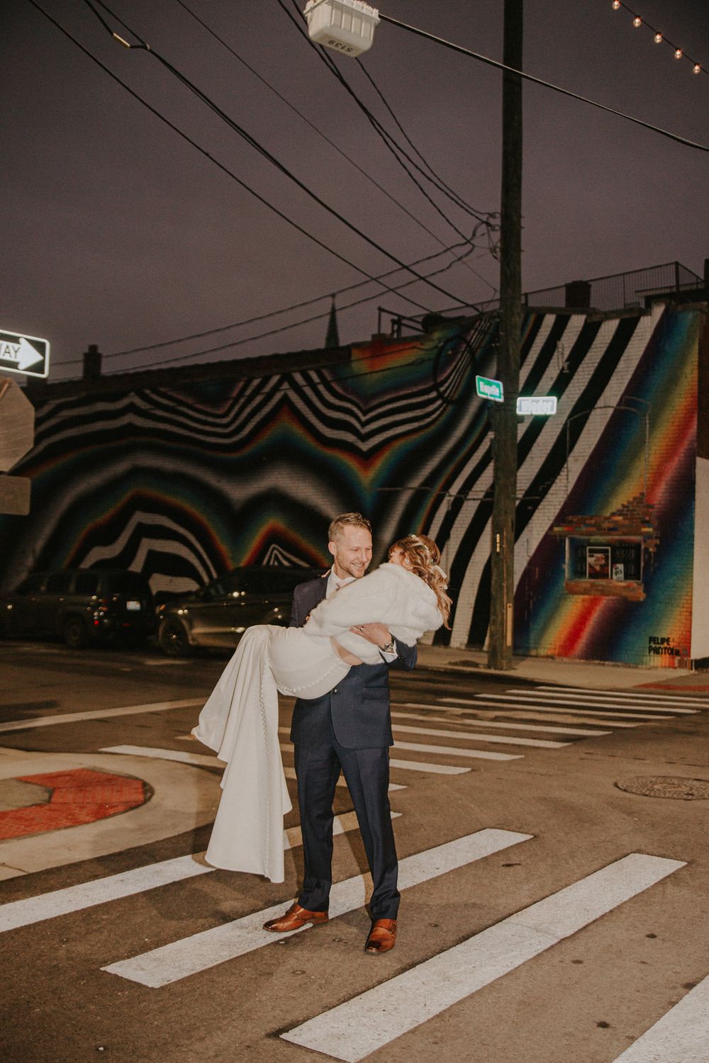 Groom carrying bride in white dress at night crosswalk with colorful rainbow mural backdrop at wedding.