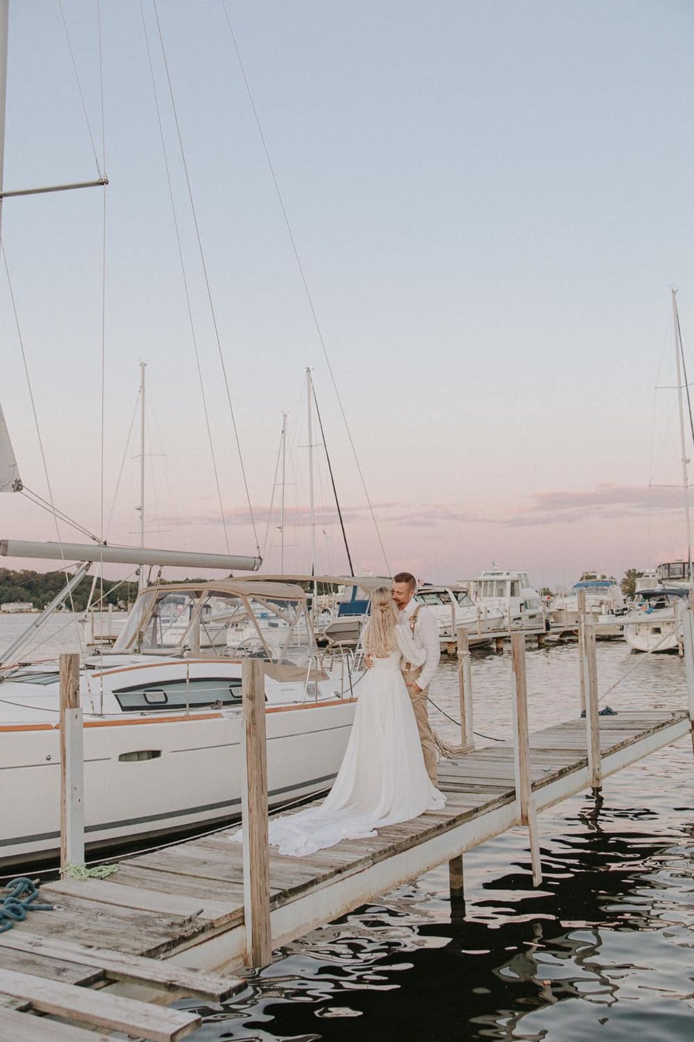 Bride and groom embracing on a wooden dock at sunset surrounded by sailboats in a marina.