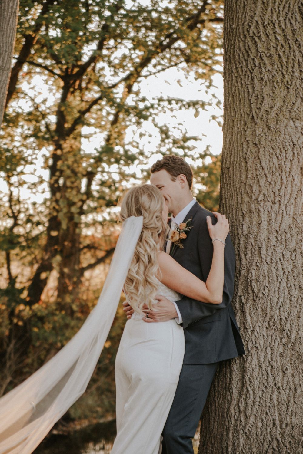 Bride and groom share a romantic kiss beside a large tree in a sunlit autumn forest, bride wearing a flowing veil.