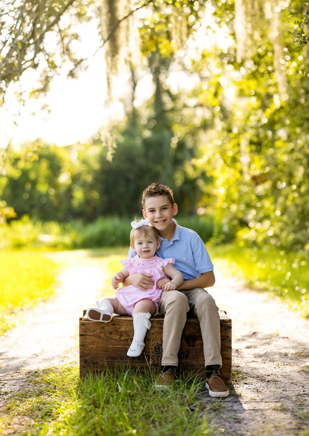 A smiling boy and baby girl in pink dress sit together on a rustic wooden crate along a sunny tree-lined path.