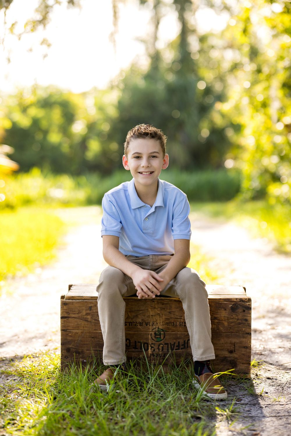 Smiling boy in blue polo shirt sitting on rustic wooden crate outdoors on sunny tree-lined path.