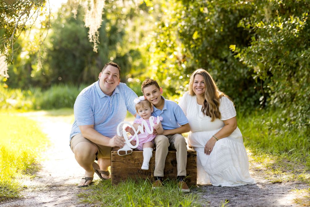 Family of four posing outdoors on a sunny path, parents flanking two young children sitting in a wooden crate.
