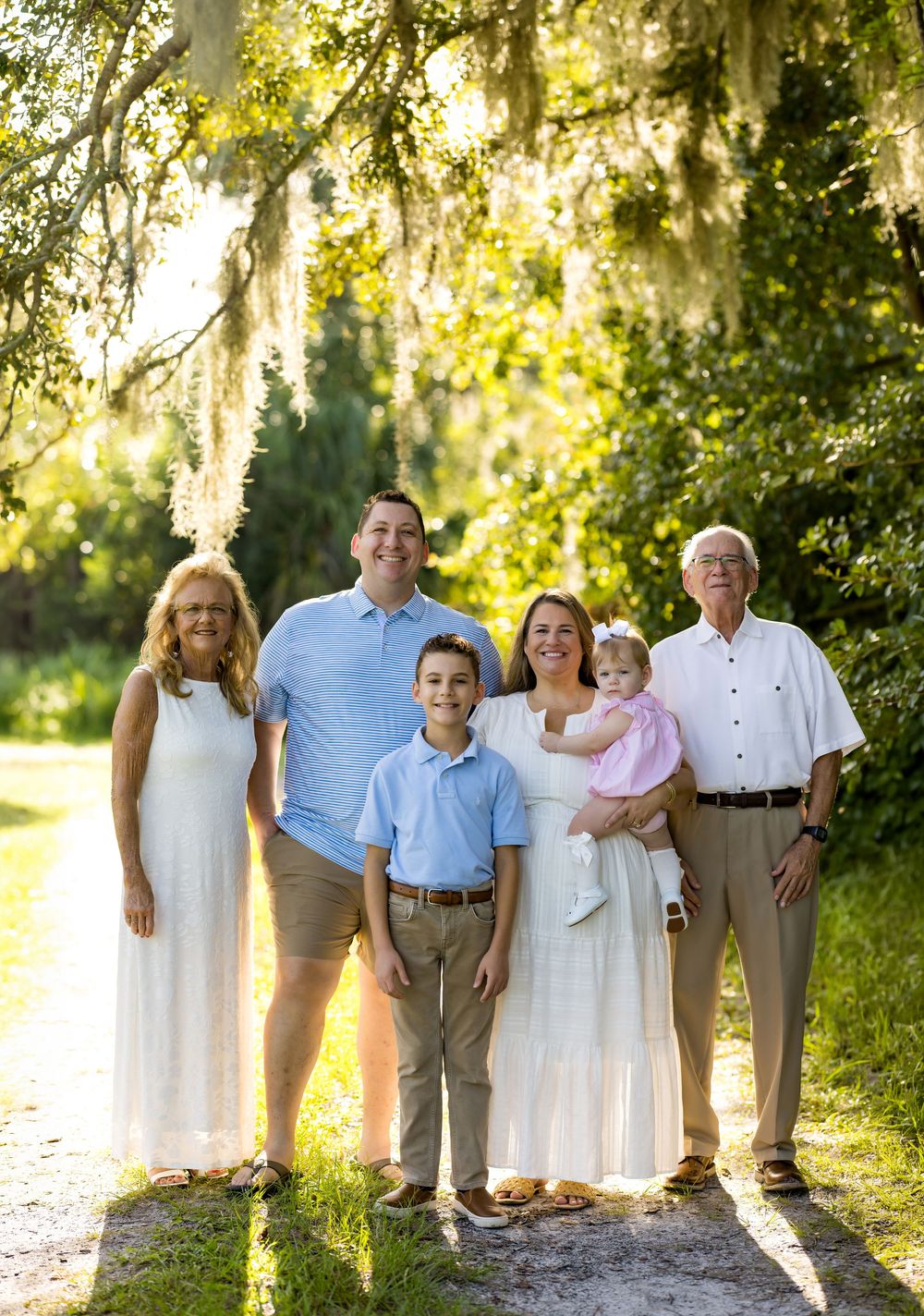 Multi-generational family of six poses on a sunlit path surrounded by lush greenery and Spanish moss-draped trees.