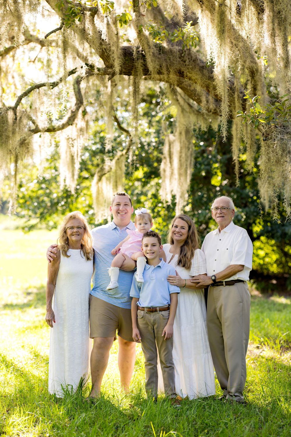 Multi-generational family portrait under Spanish moss-draped oak tree on sunny green lawn.