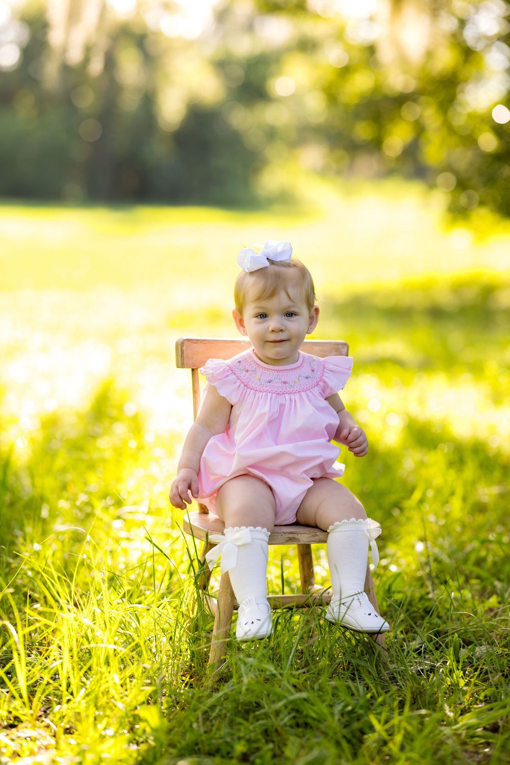 Adorable baby girl in pink smocked romper and white bow sits on wooden chair in sunny green meadow.