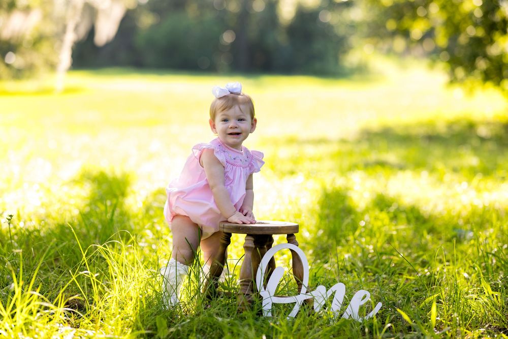 Baby girl in pink romper sits on small wooden bench in sunny green field beside decorative One sign for first birthday.
