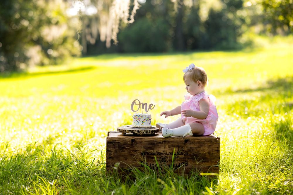 A baby girl in pink sits on a wooden crate with a first birthday cake topper reading One in a sunny green field.