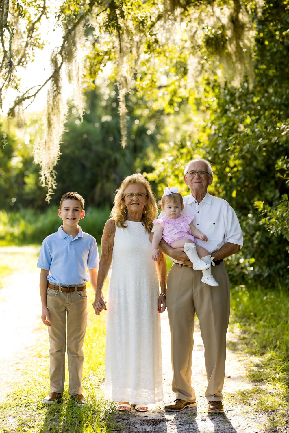 Grandparents posing outdoors with grandson and baby granddaughter under Spanish moss-draped trees in golden sunlight.