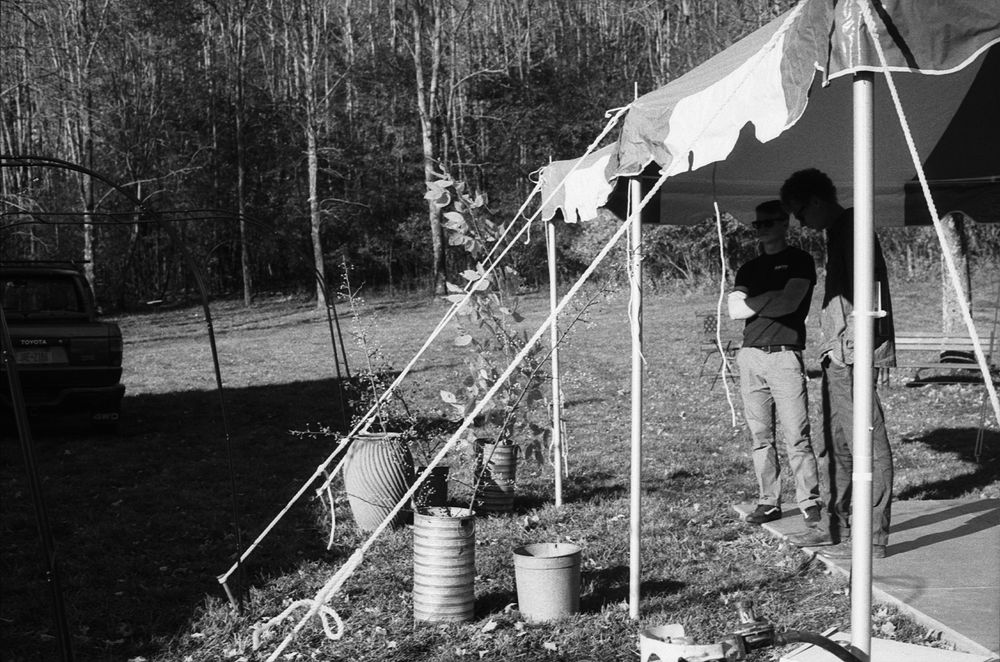 Black and white photo of a person standing under a canvas tent outdoors near trees with buckets and supplies.