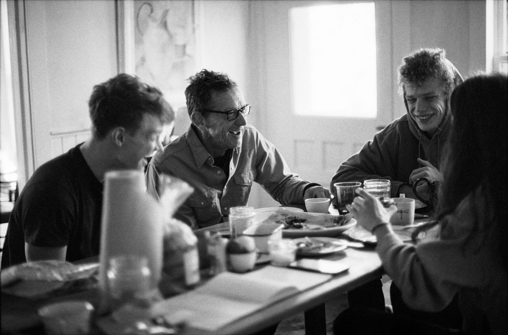 Black and white photo of four people sharing a meal at a table, engaged in lively conversation in a bright room.