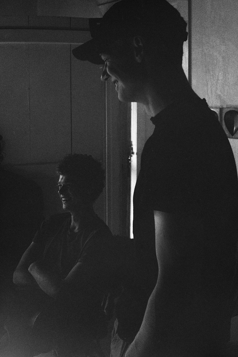 Black and white photo of two men in a dimly lit room, one wearing a cap, with dramatic light contrast.