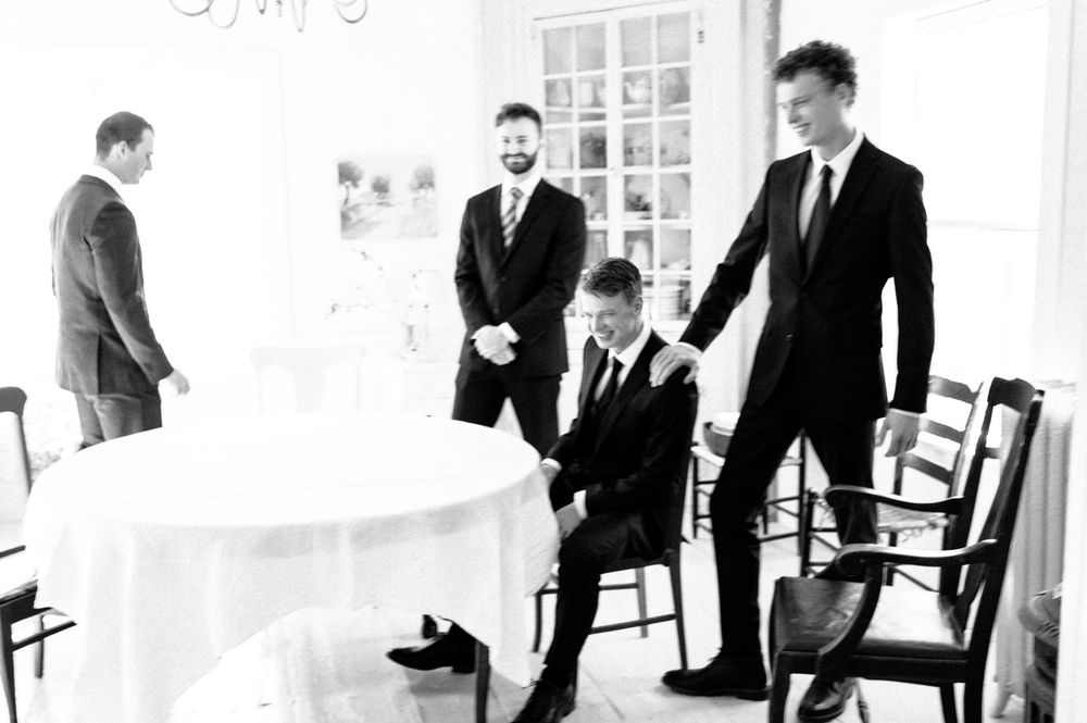 Black and white photo of groomsmen in suits preparing together in an elegant white room before a wedding ceremony.