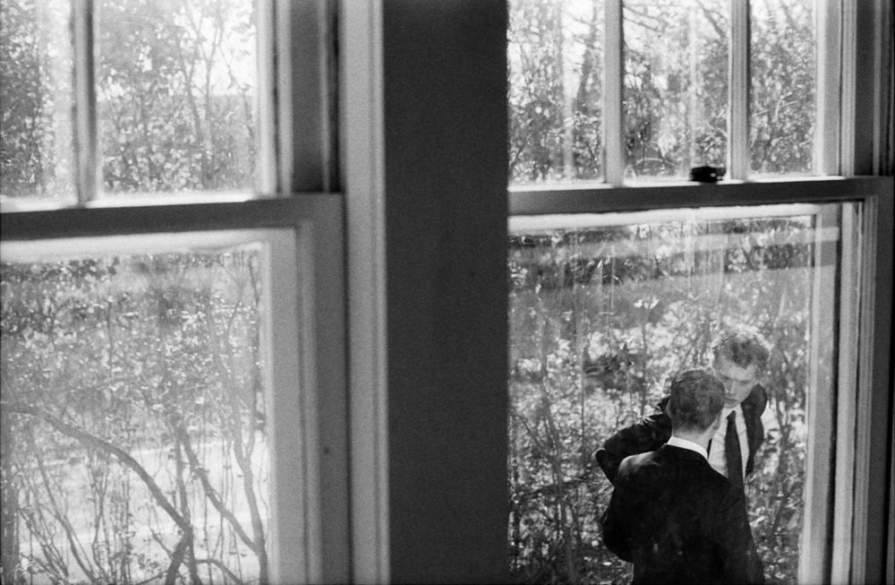 Black and white photo of a person looking through binoculars by a window with trees visible outside.
