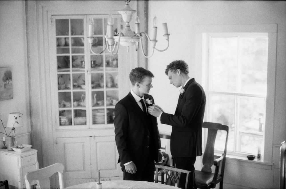 Groom getting boutonniere pinned by groomsman in black suits inside elegant room, black and white wedding photo.