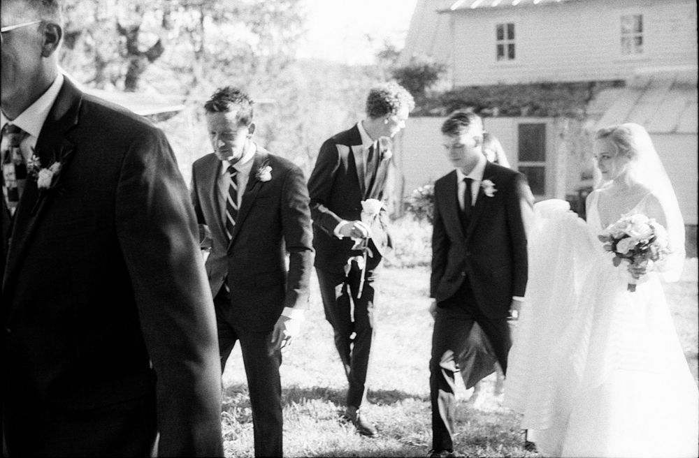 Black and white wedding photo of bride and groom with groomsmen walking outdoors near a white building.
