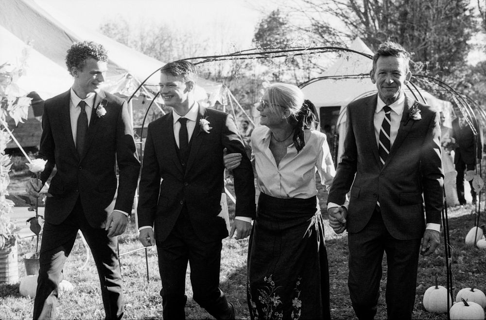 Wedding party of four walking outdoors laughing in black and white photo, men in suits and woman in blouse.