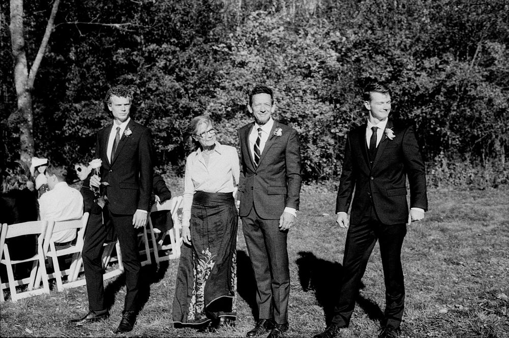 Black and white photo of wedding party walking outdoors near white chairs and trees.