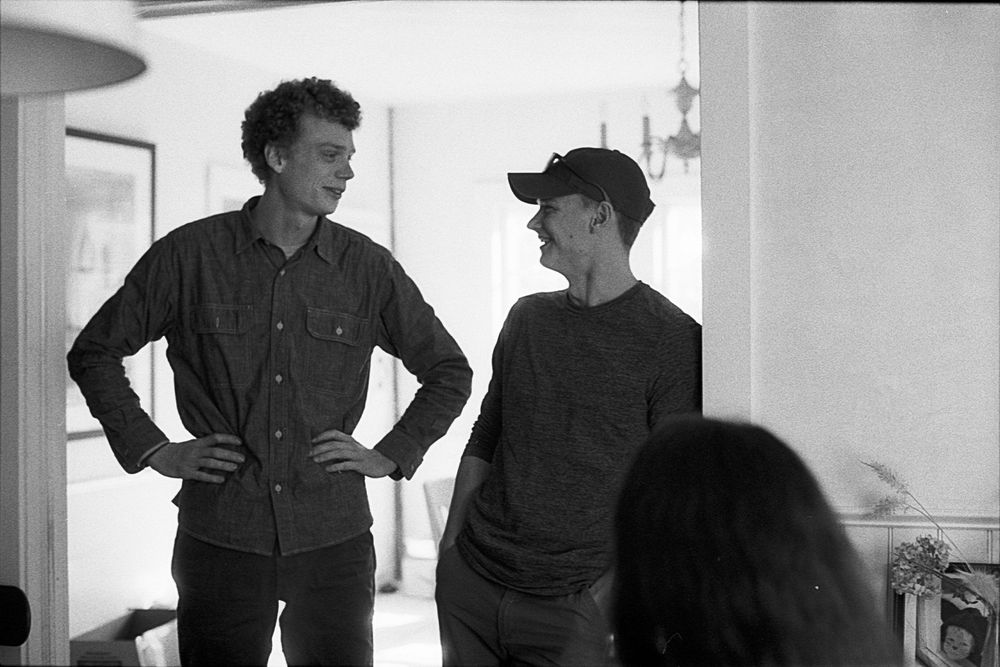Two men stand talking indoors in a black and white photo, one tall in a denim shirt, the other in a cap.