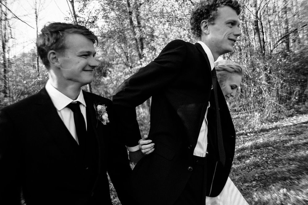 Black and white wedding photo of groom with groomsmen walking outdoors among trees in formal suits.