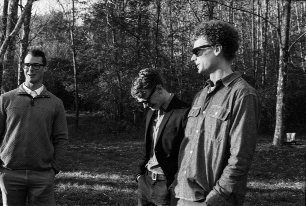 Three men standing outdoors in a wooded area, captured in black and white photography.