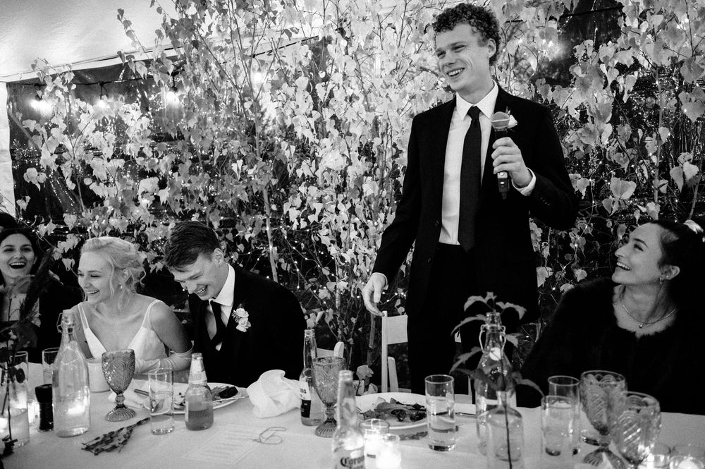 Black and white wedding reception photo of a man giving a toast while guests smile and laugh at a floral-decorated table.