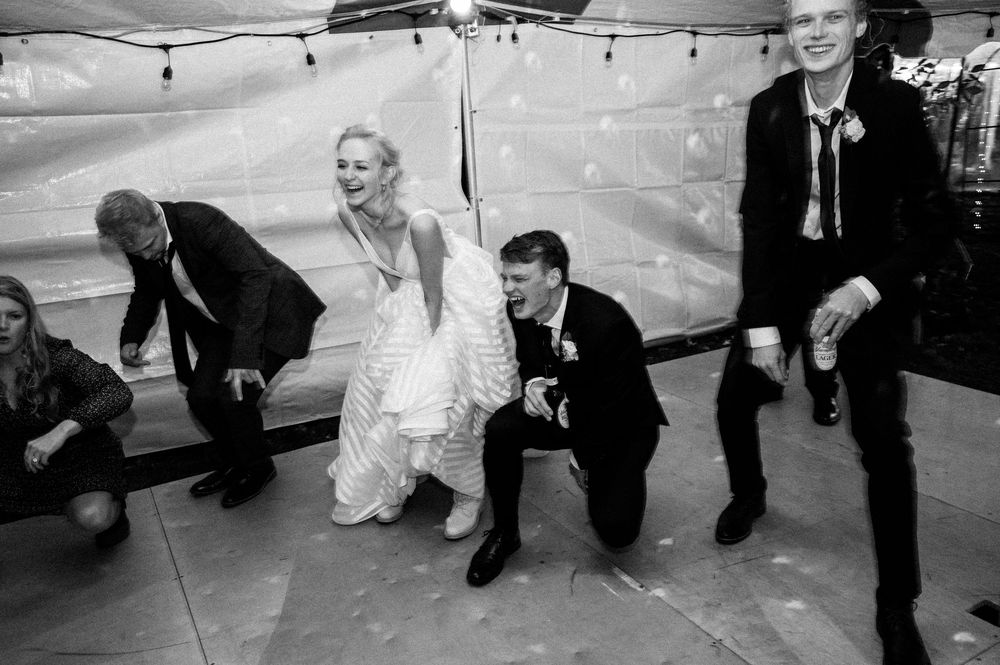 Black and white wedding photo of bride and groom dancing with guests under a tent at a lively reception.