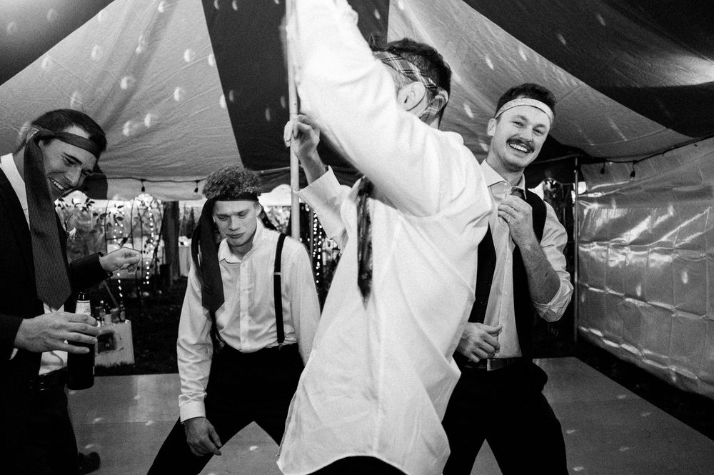 Groomsmen dancing and celebrating at wedding reception under festive tent in black and white photo.