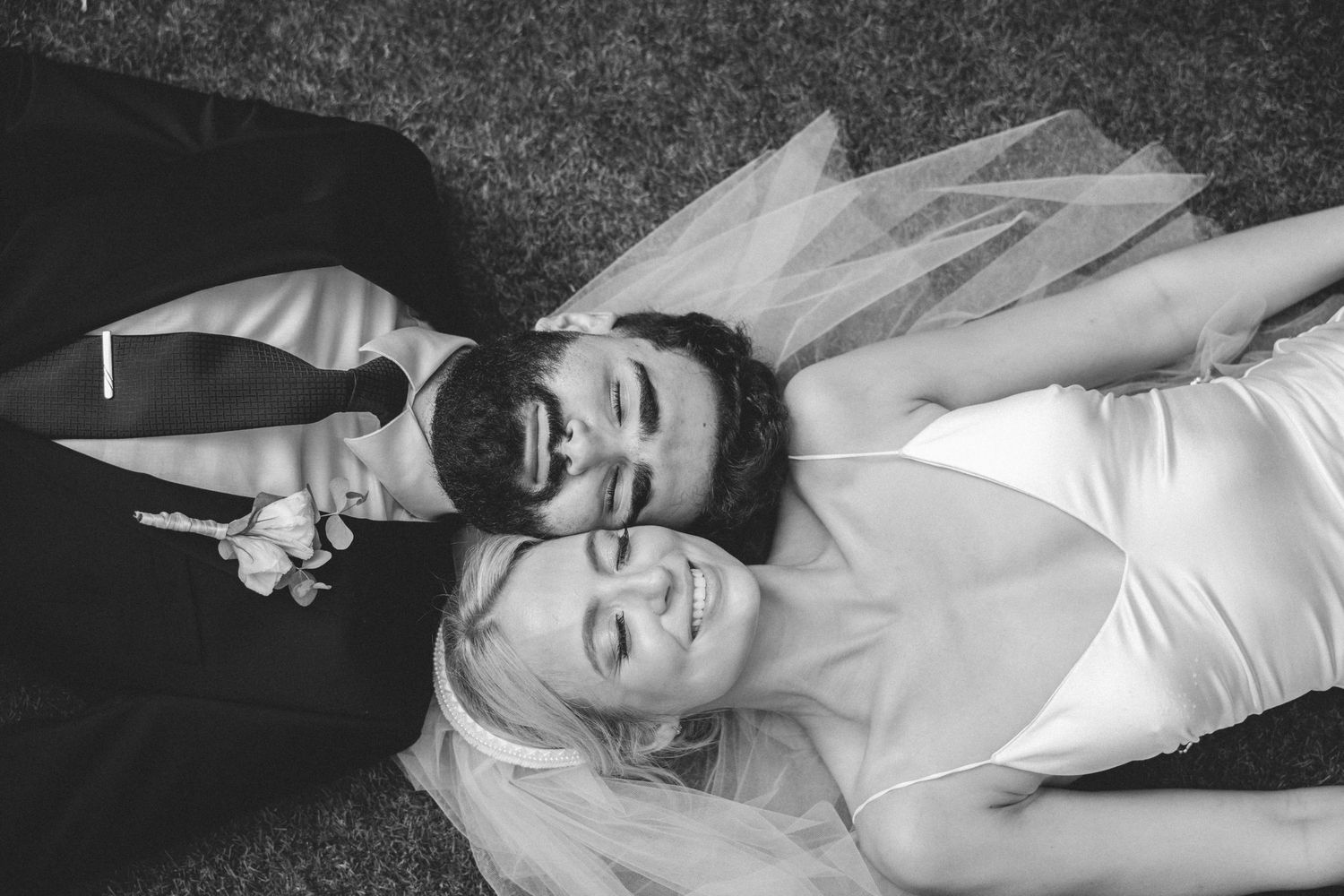 Black and white photo of smiling bride and groom lying on grass, holding hands on their wedding day. Couple's testimonial.