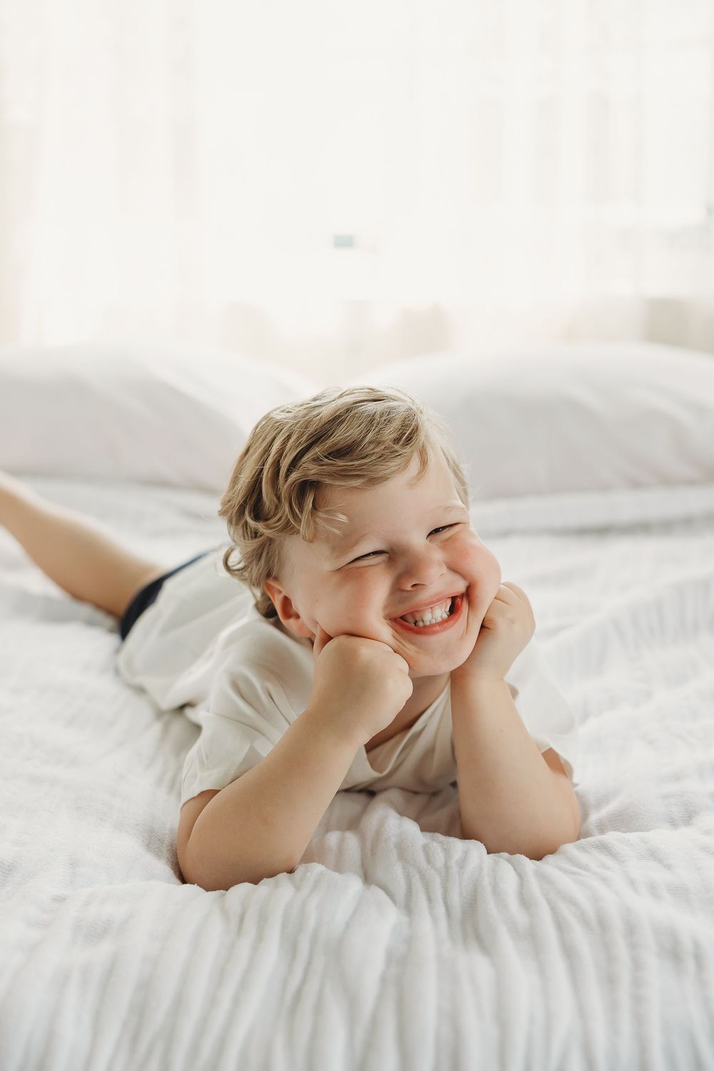 Happy toddler boy lying on white bed, smiling with hands on cheeks in bright bedroom.