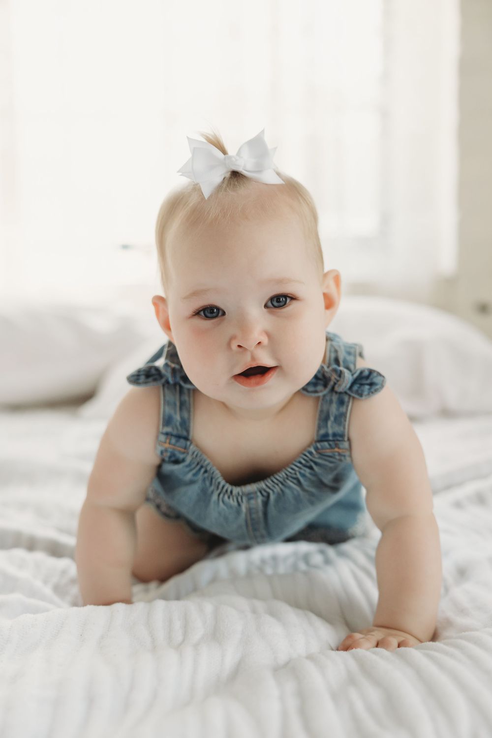 Adorable baby girl in denim overalls and white bow crawling on white bed.