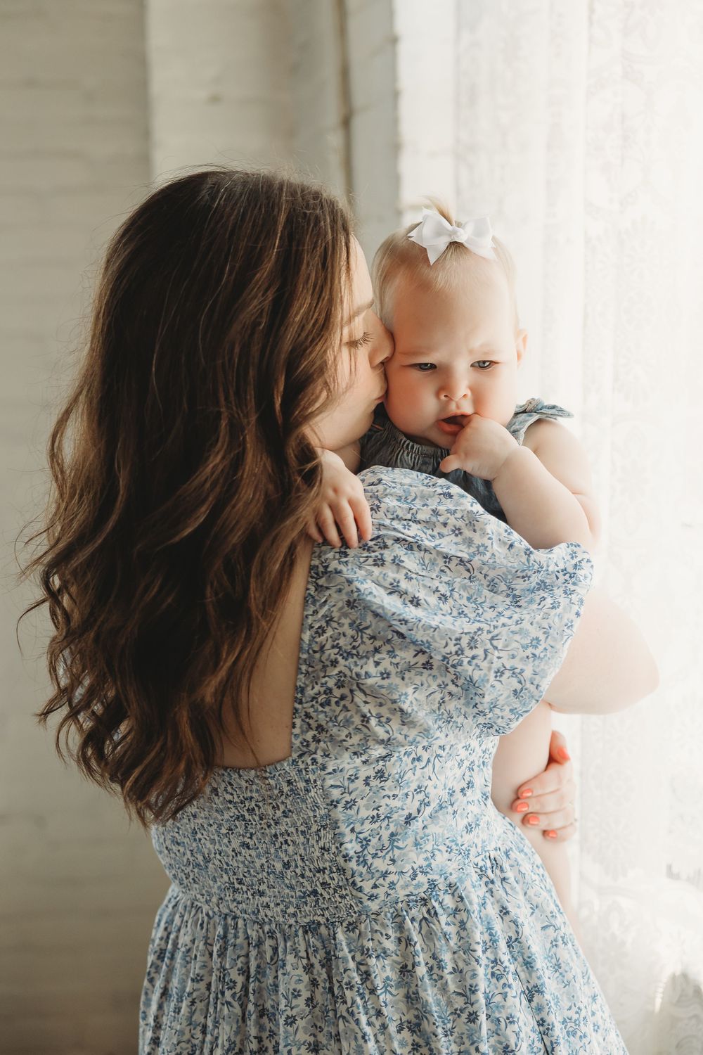 A mother in a floral dress kisses her baby girl with a white bow near a bright window.