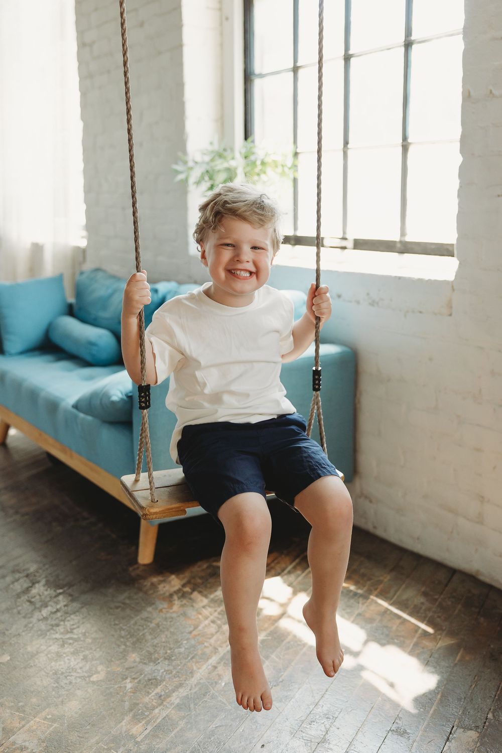 Happy young boy sitting on an indoor rope swing, wearing white t-shirt and navy shorts, smiling joyfully.