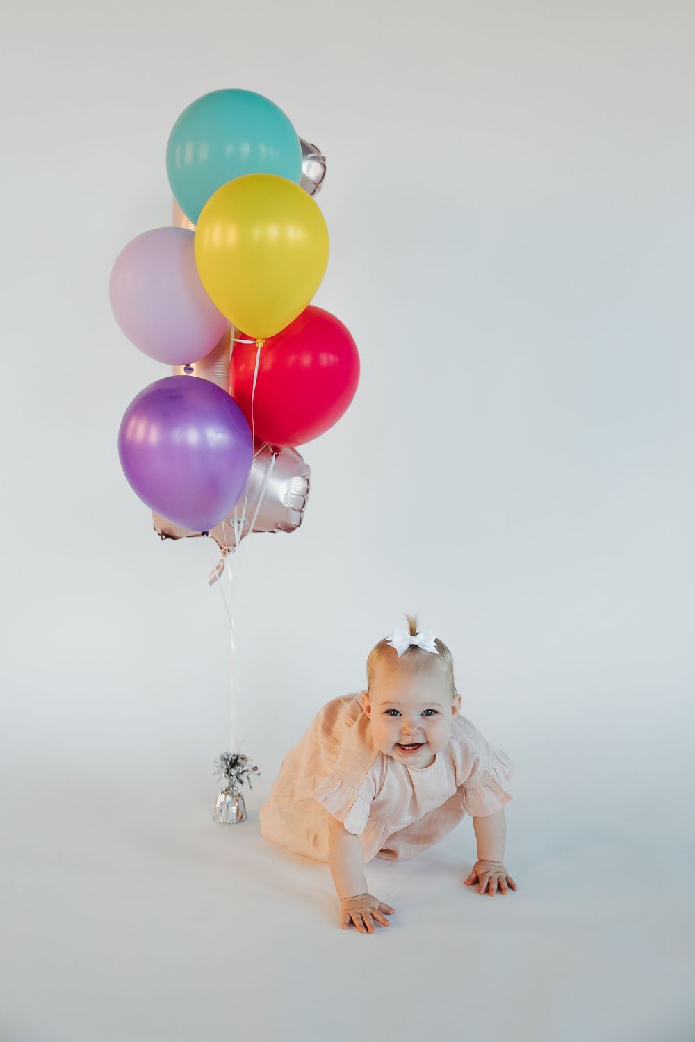 Happy baby girl in pink tutu crawls on white background beside colorful birthday balloons bundle.