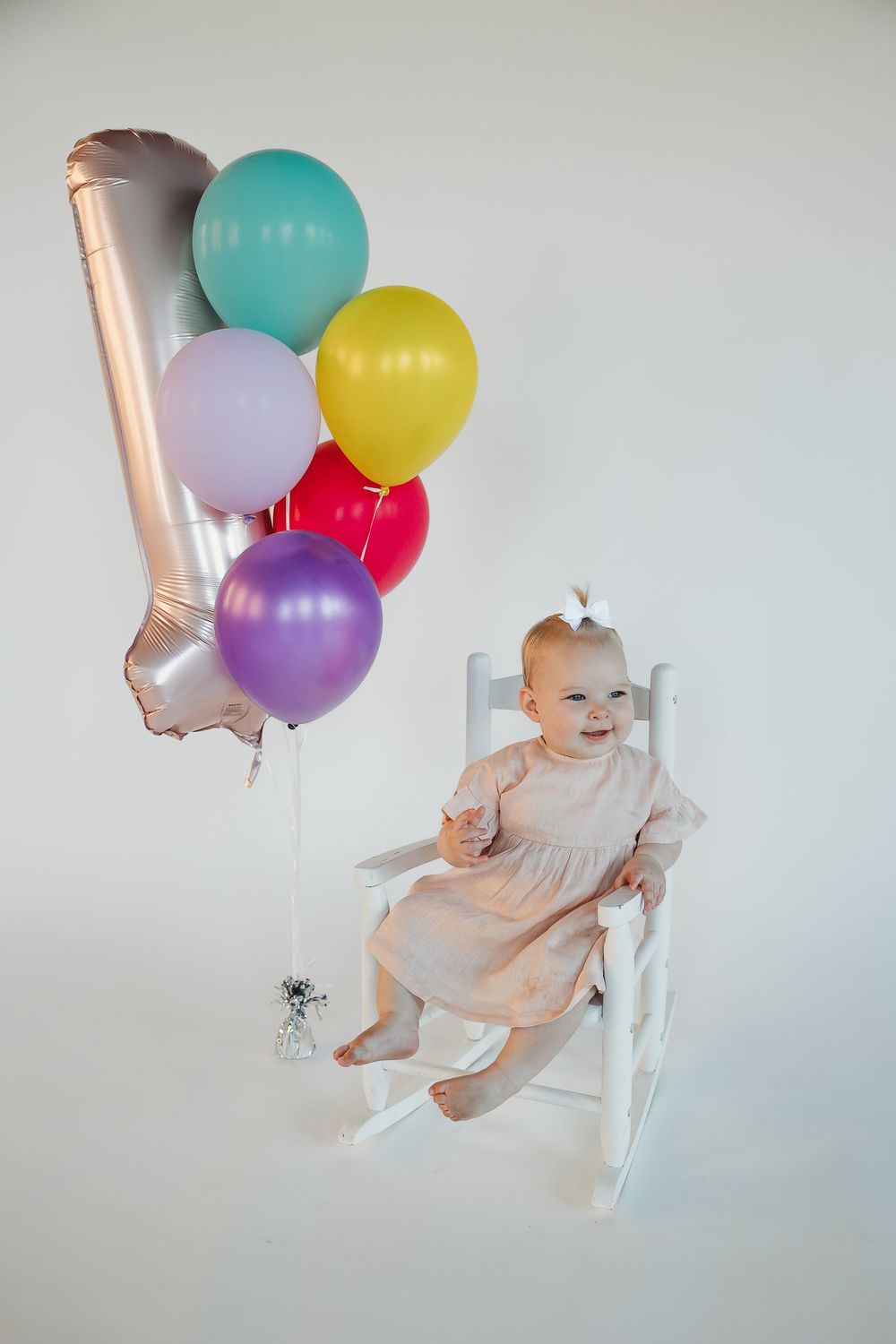 Baby girl in pink dress sits in white rocking chair smiling, surrounded by colorful balloons for her 1st birthday.