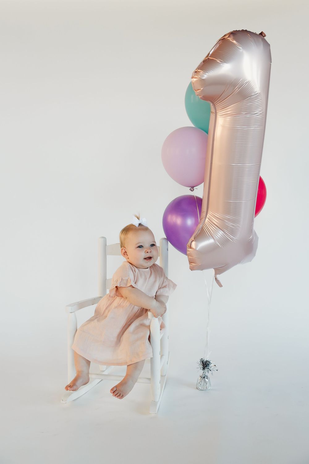 Baby girl in pink dress sits in white rocking chair beside number 1 balloon and colorful balloons for first birthday.