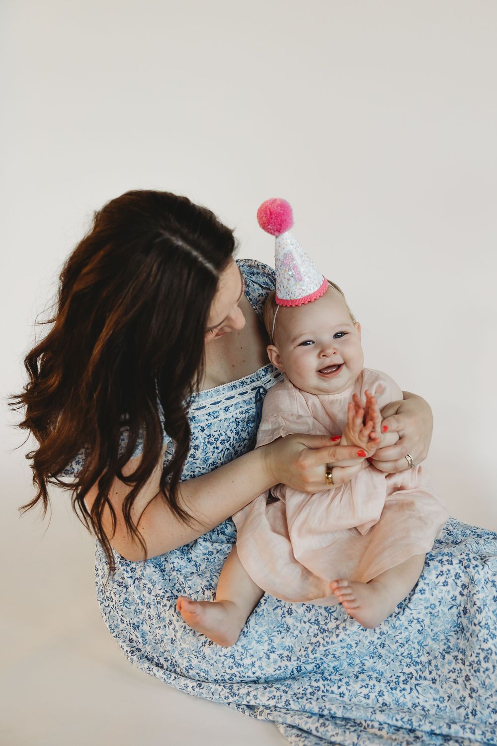 A mother in a blue dress holds her smiling baby wearing a pink birthday hat, celebrating a first birthday.