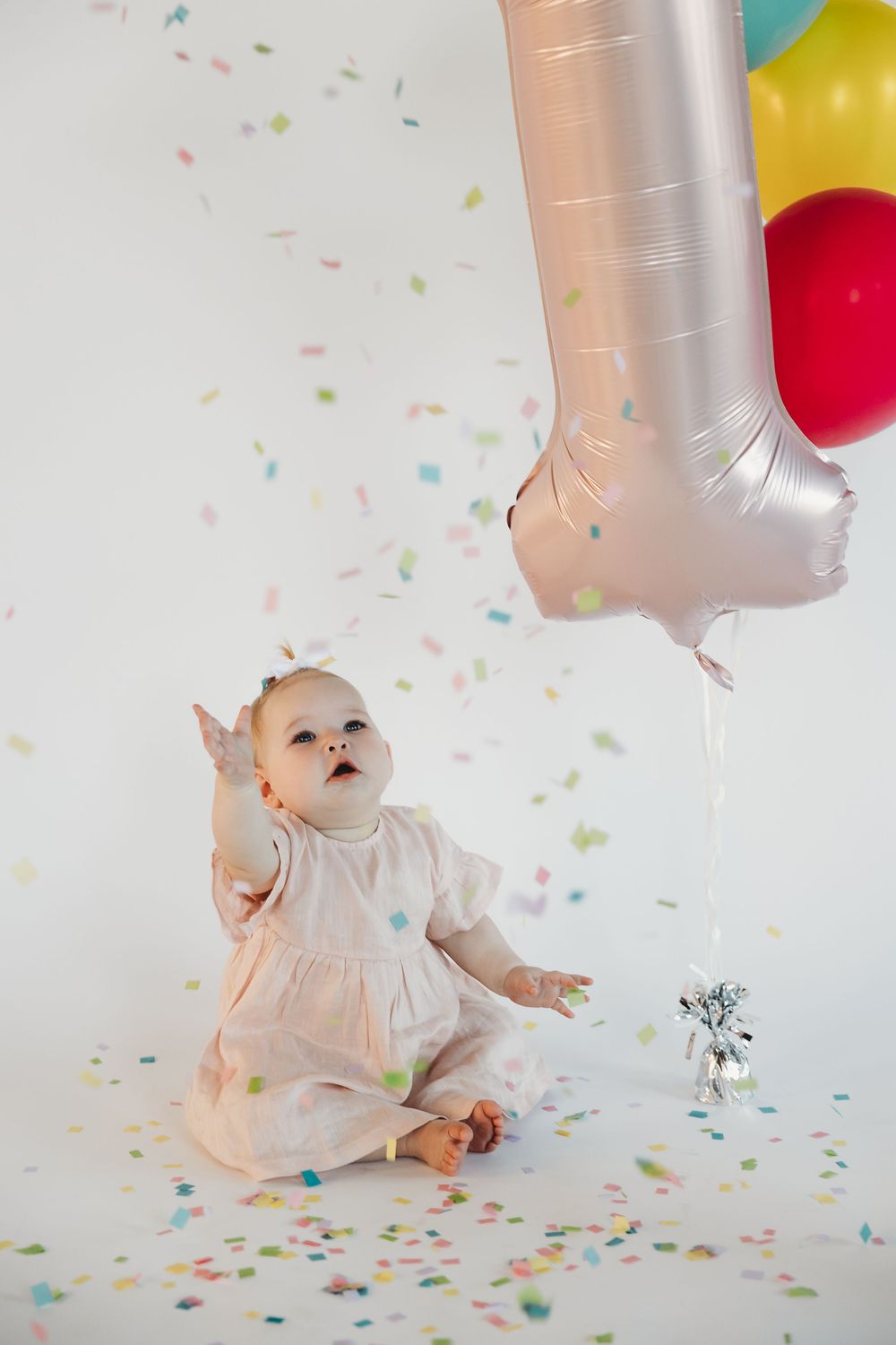 Baby girl in white dress sits among falling confetti beside a number one balloon on her first birthday.