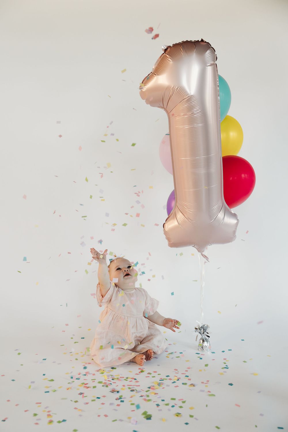 Baby girl in white dress celebrates first birthday with number one balloon and colorful confetti on white background.
