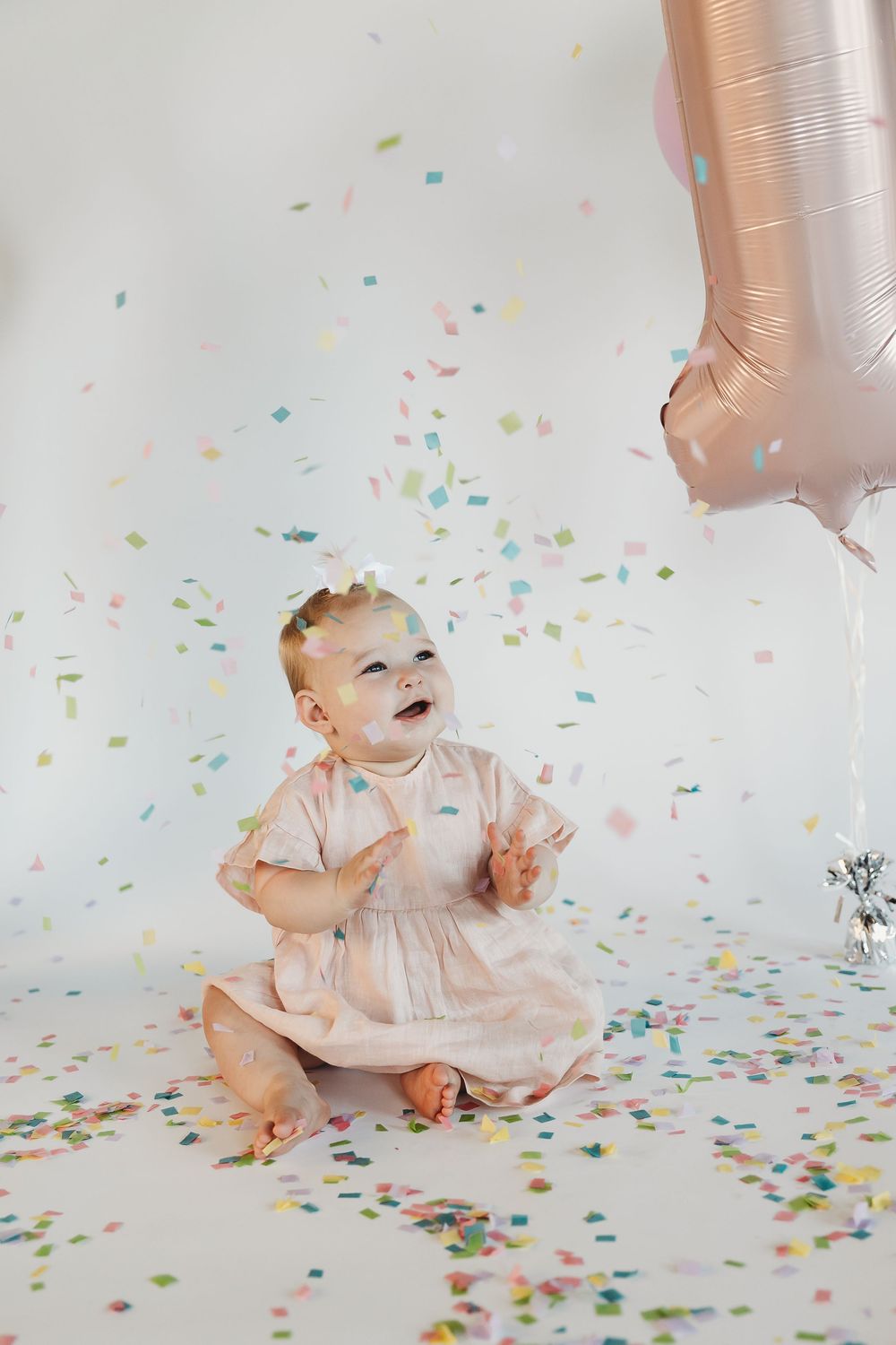 Happy baby girl in pink dress celebrating first birthday with colorful confetti and number 1 balloon.