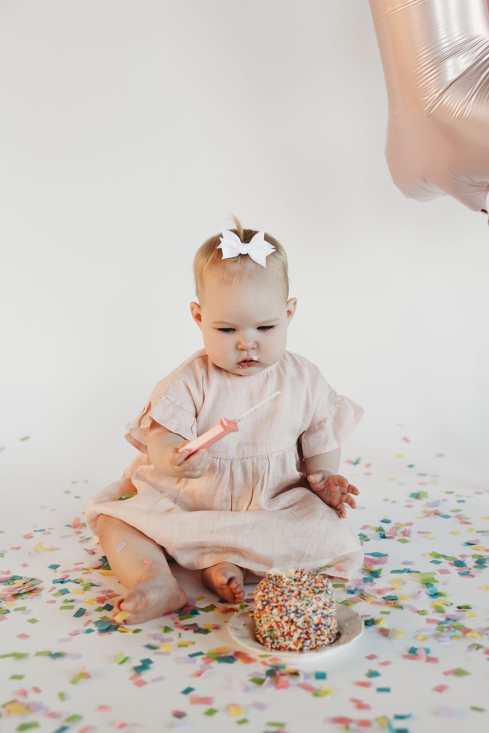 Baby girl in pink dress sits on white floor with birthday cake, candle, and colorful confetti during first birthday smash.
