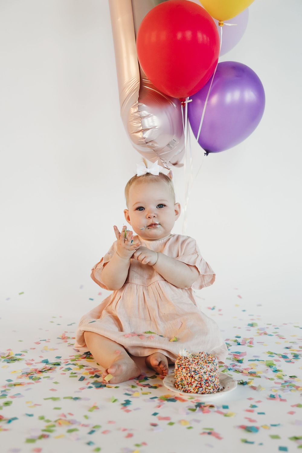 Baby girl in pink dress celebrates first birthday with colorful balloons and smash cake on white background.