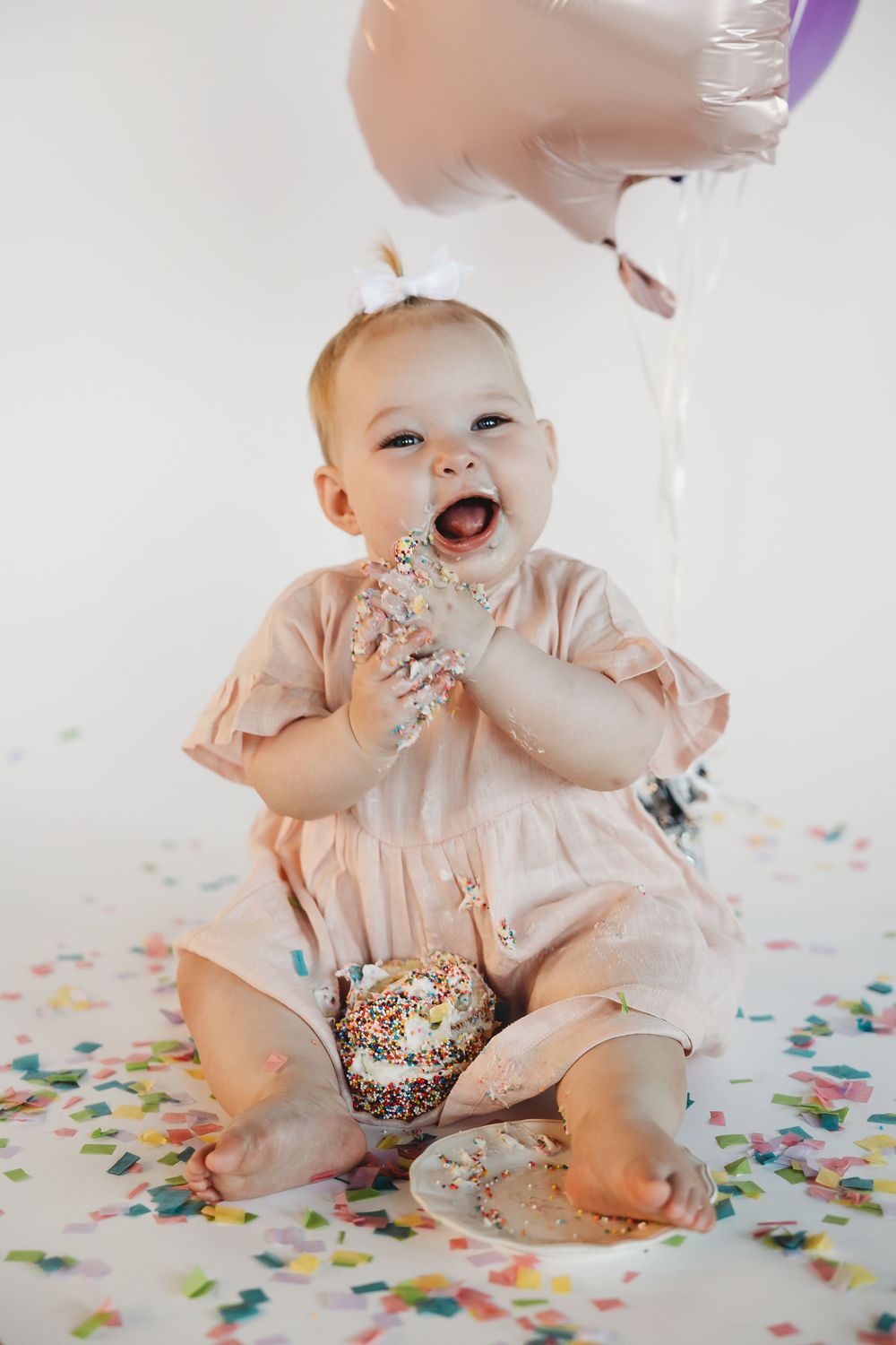 Cute baby girl in pink dress eating birthday cake with sprinkles, surrounded by colorful confetti and balloons.