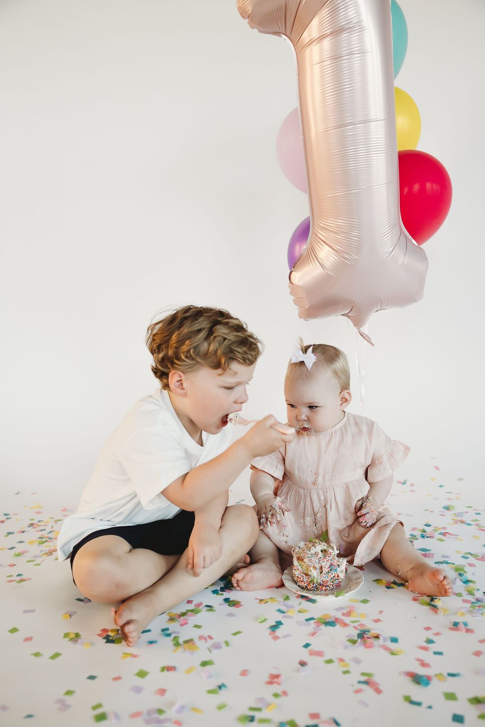 A young boy feeds cake to a baby girl on her 1st birthday, surrounded by colorful balloons and confetti.